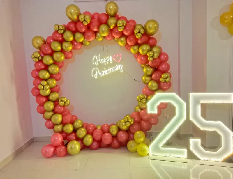 Coral and gold balloon ring with a neon Happy Anniversary sign and a lit 25 standing beside it.