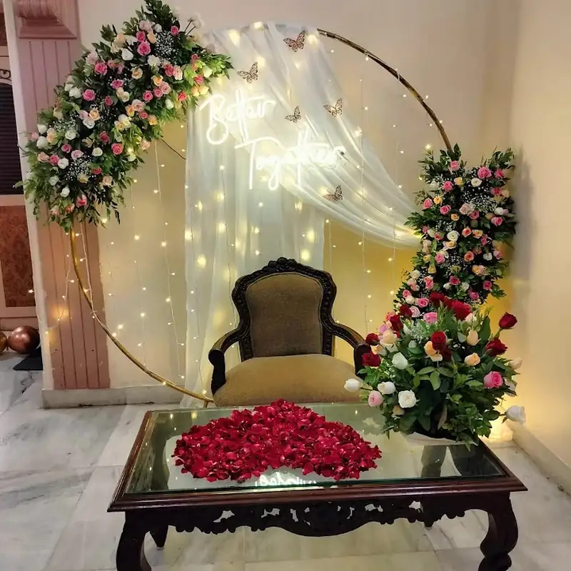 Flower hoop with a lit drape, a carved chair and red rose petals arranged on a low table.