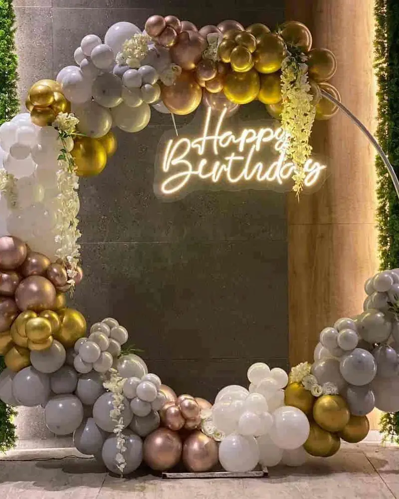 A ring of chrome and gold balloons around a neon Happy Birthday sign, set against a green hedge wall.