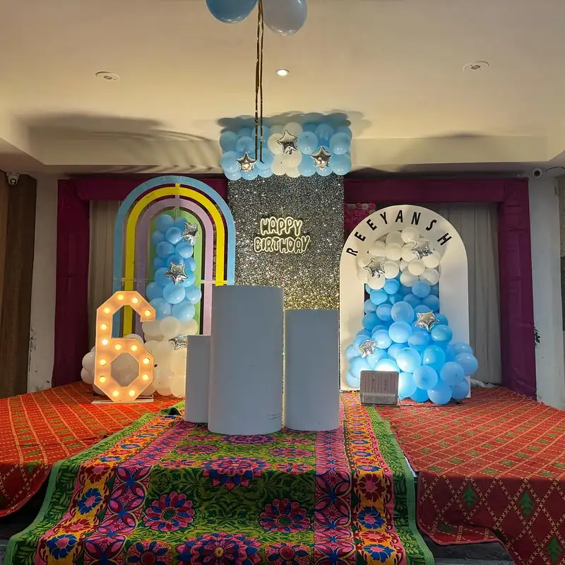 Blue and white balloon backdrop with a rainbow arch and a name in bulb letters in a banquet hall.